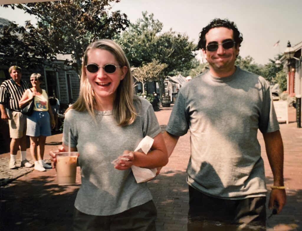 Two smiling individuals walk together on a sunny street, holding drinks and snacks. Background figures are partially visible.