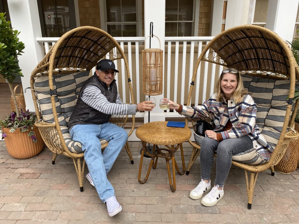Two people toast with drinks while sitting in large woven chairs, surrounded by plants and a cozy setting.