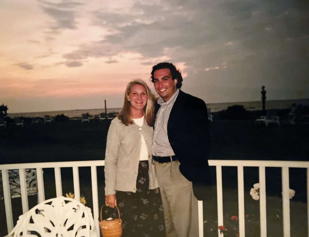 A woman and a man stand together on a balcony, smiling against a sunset backdrop over the ocean.