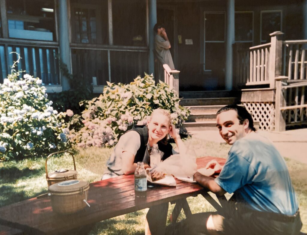 Two people sit at a picnic table, smiling and enjoying food, with flowers and a building in the background.