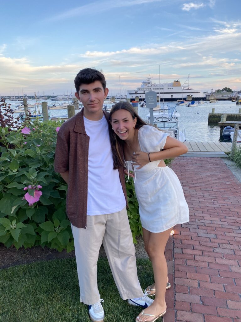 Two young adults pose together outdoors, smiling, with a marina and boats in the background.