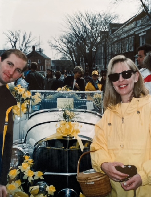 Two people pose beside a decorated vintage car with yellow flowers at a crowded outdoor event.