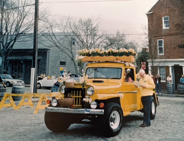 A decorated yellow truck with flowers drives past people during a parade. A man in a yellow shirt stands beside it.