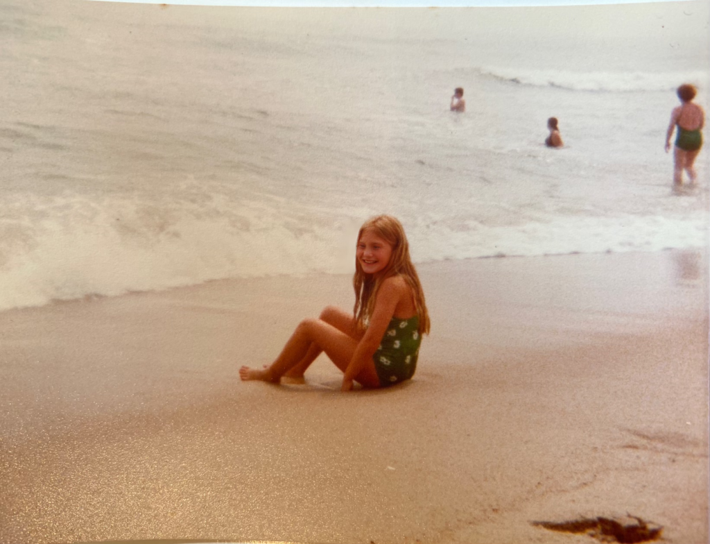 A girl sits on the beach, smiling, with waves in the background and other children playing in the water.