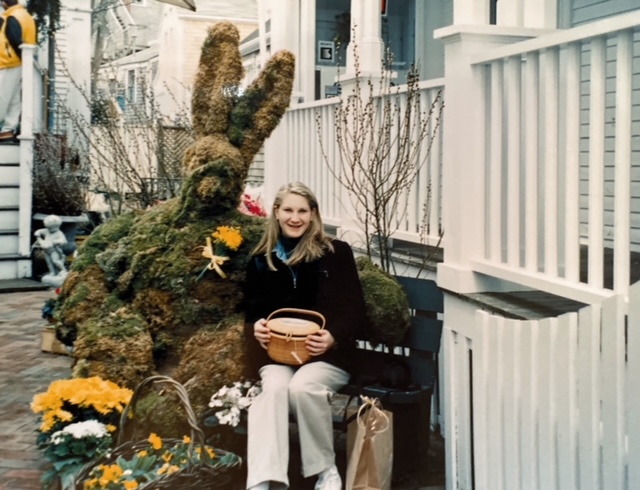 A woman sits on a bench beside a large moss bunny sculpture, surrounded by flowers and plants.