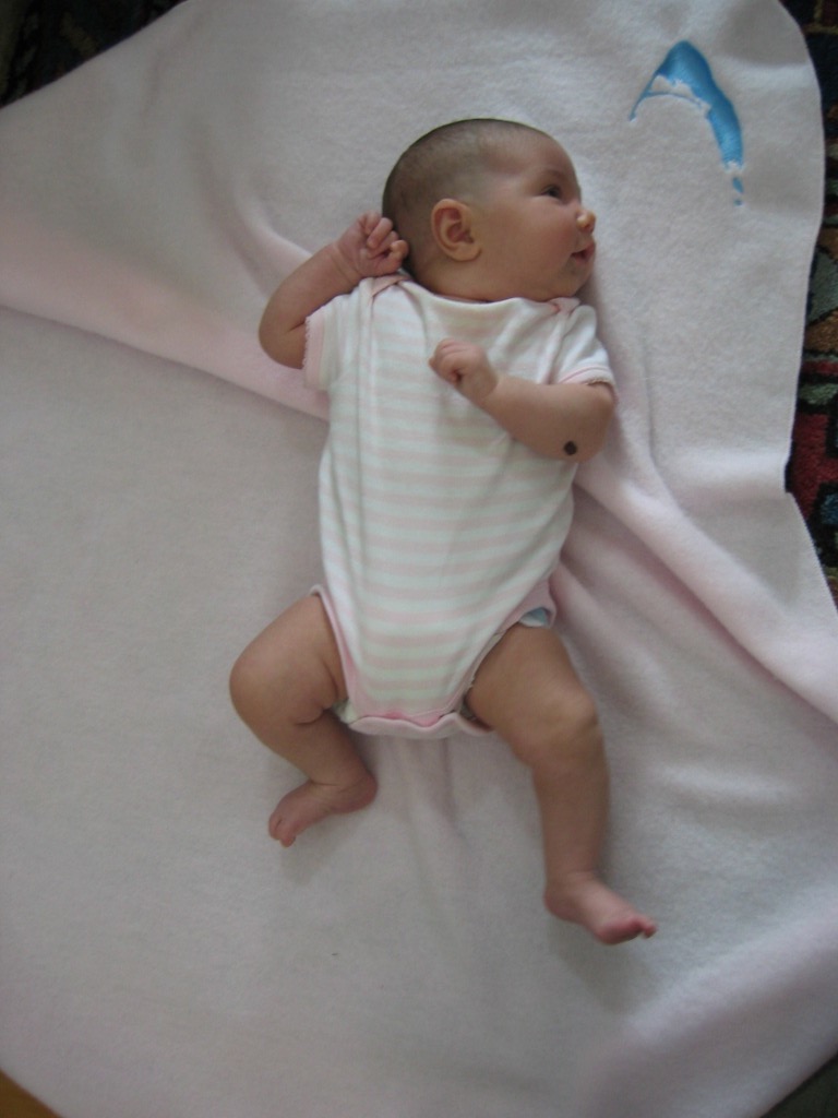 A baby lies on a pink blanket, looking to the side with one arm raised.