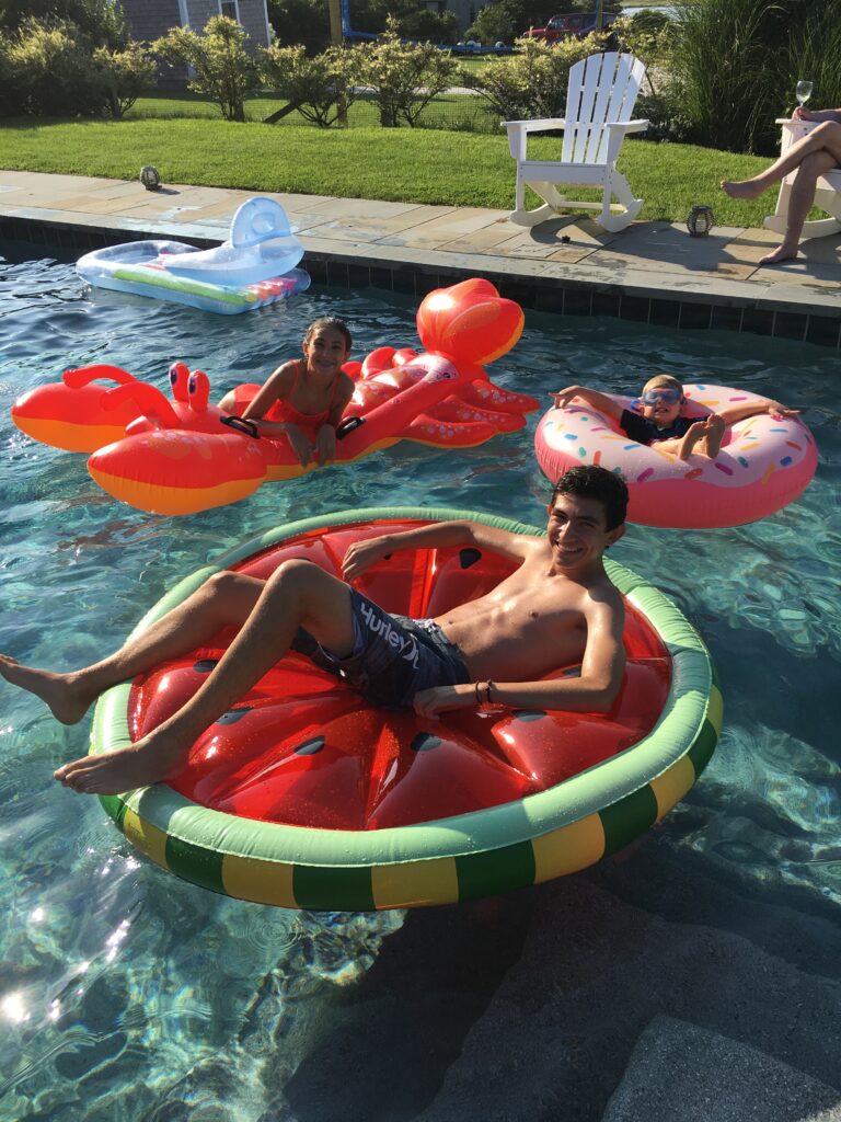 Three children float on colorful pool inflatables; a watermelon, lobster, and donut, enjoying a sunny day.