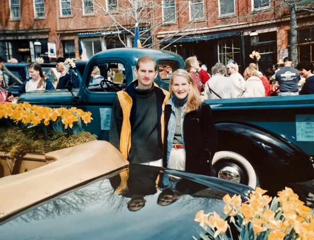 A man and woman pose in front of a vintage truck, surrounded by a crowd and blooming flowers.
