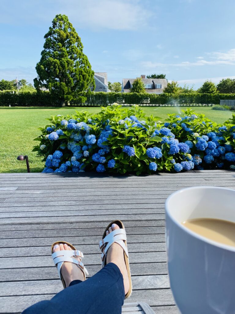 Person's feet in white sandals rest on a wooden deck, with a coffee cup and blue hydrangeas in the background.