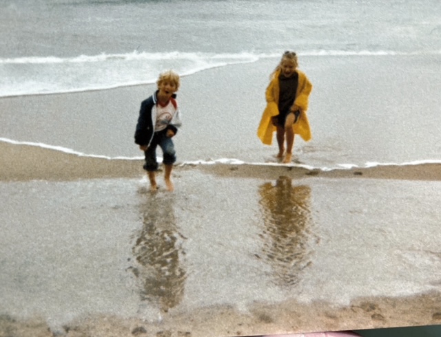 Two children play at the beach, wading in shallow water and reflecting in the wet sand.