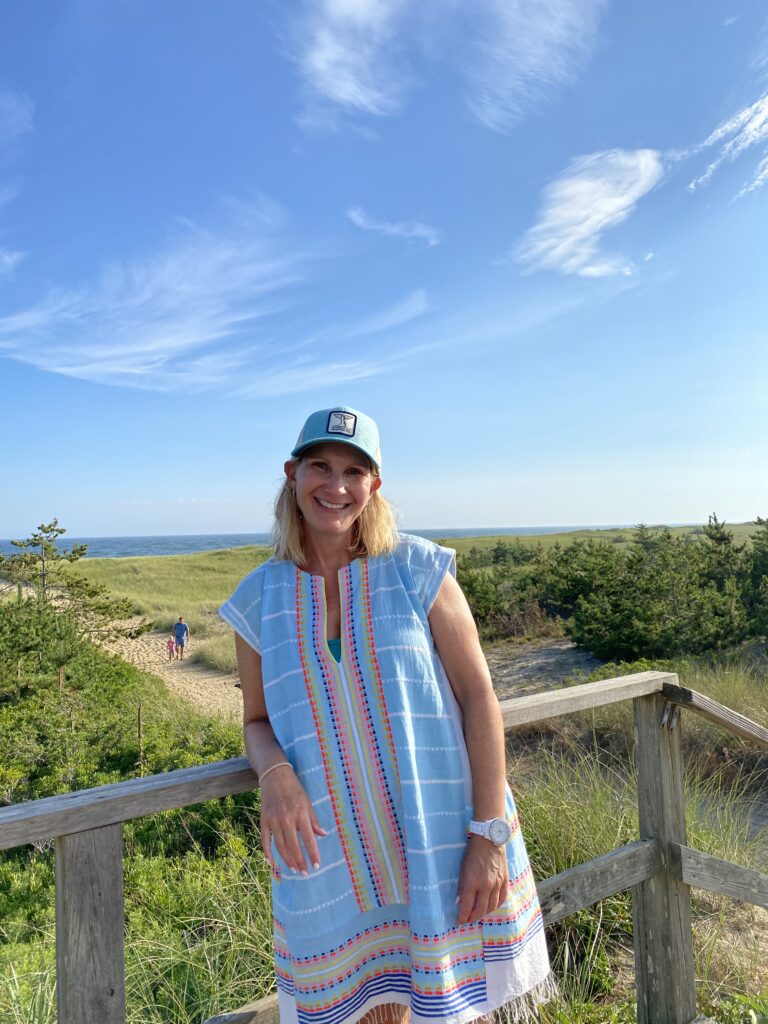 A woman in a blue dress stands on a wooden deck, smiling with a beach and greenery in the background.