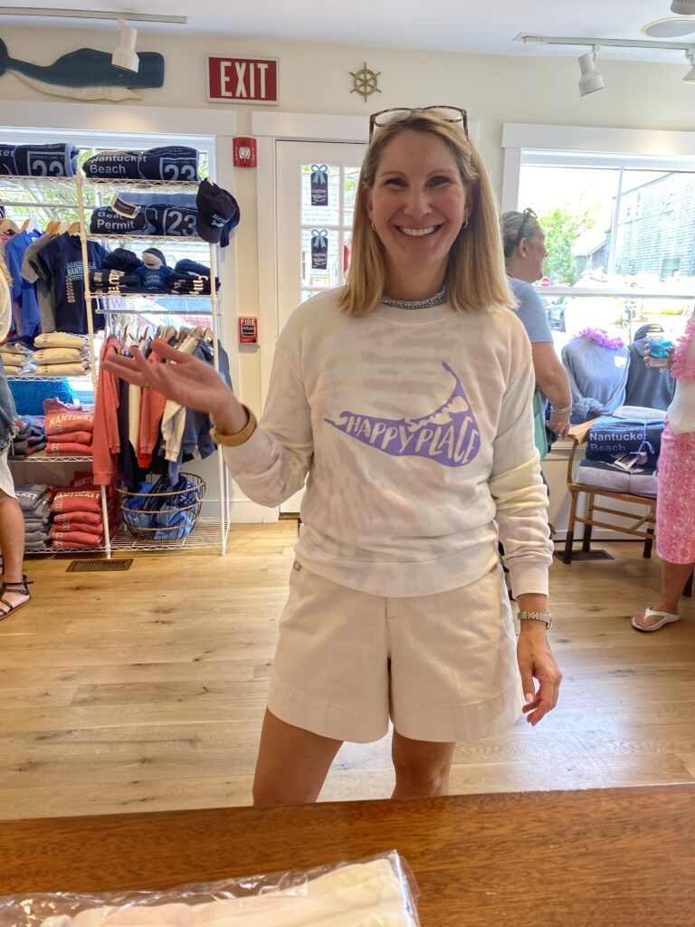 A woman in a "Happy Place" sweatshirt smiles and gestures in a clothing store filled with merchandise.