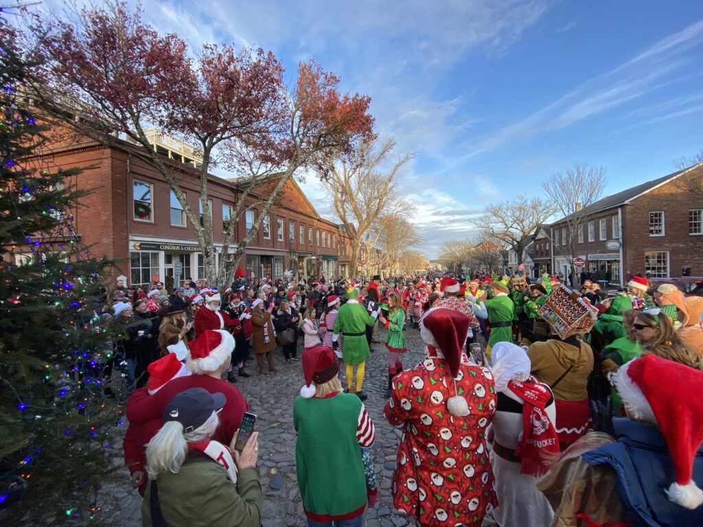Crowd in festive attire gathers on a cobblestone street, celebrating with decorations and holiday spirit.