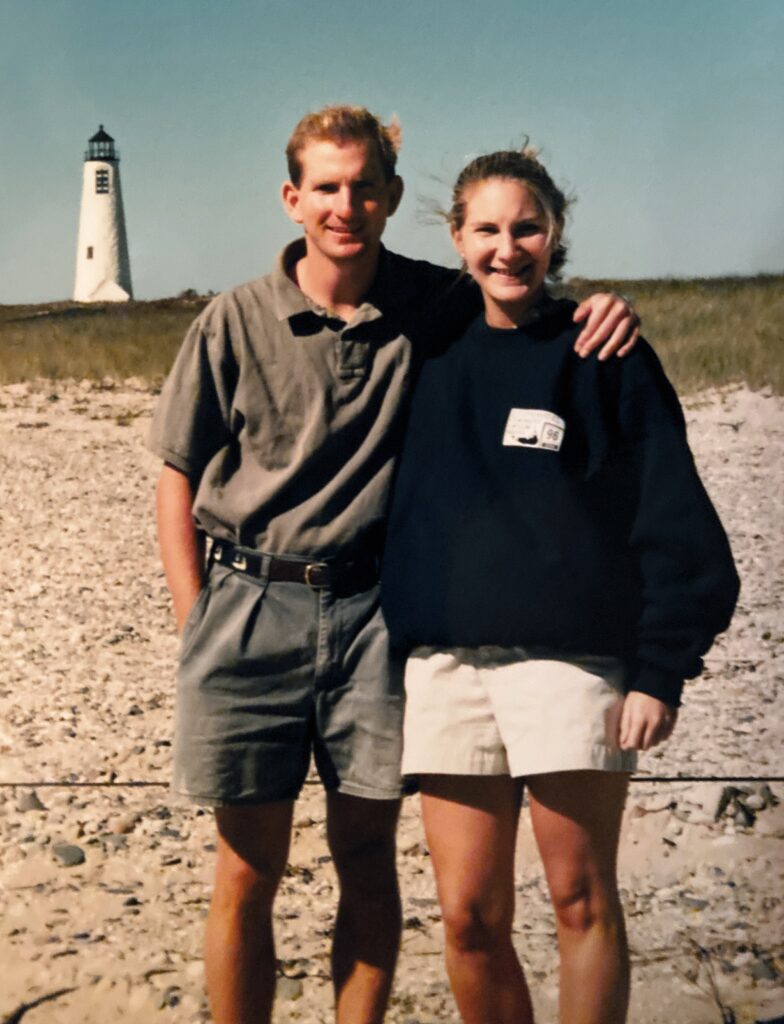 Two people stand together on a beach, smiling, with a lighthouse in the background.