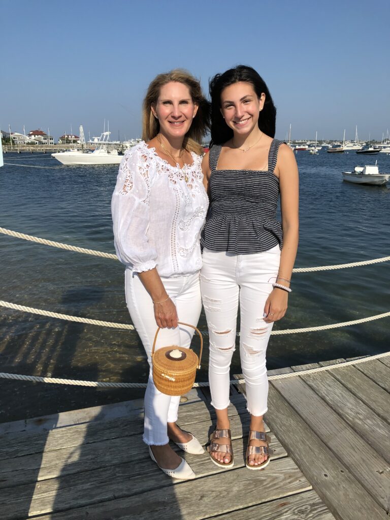 Two women stand on a dock by the water, smiling. One holds a small woven basket. Boats are visible in the background.