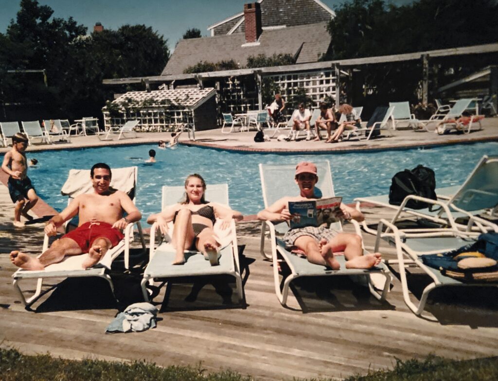 Three people relax on lounge chairs by a swimming pool, while others swim and socialize in the background.