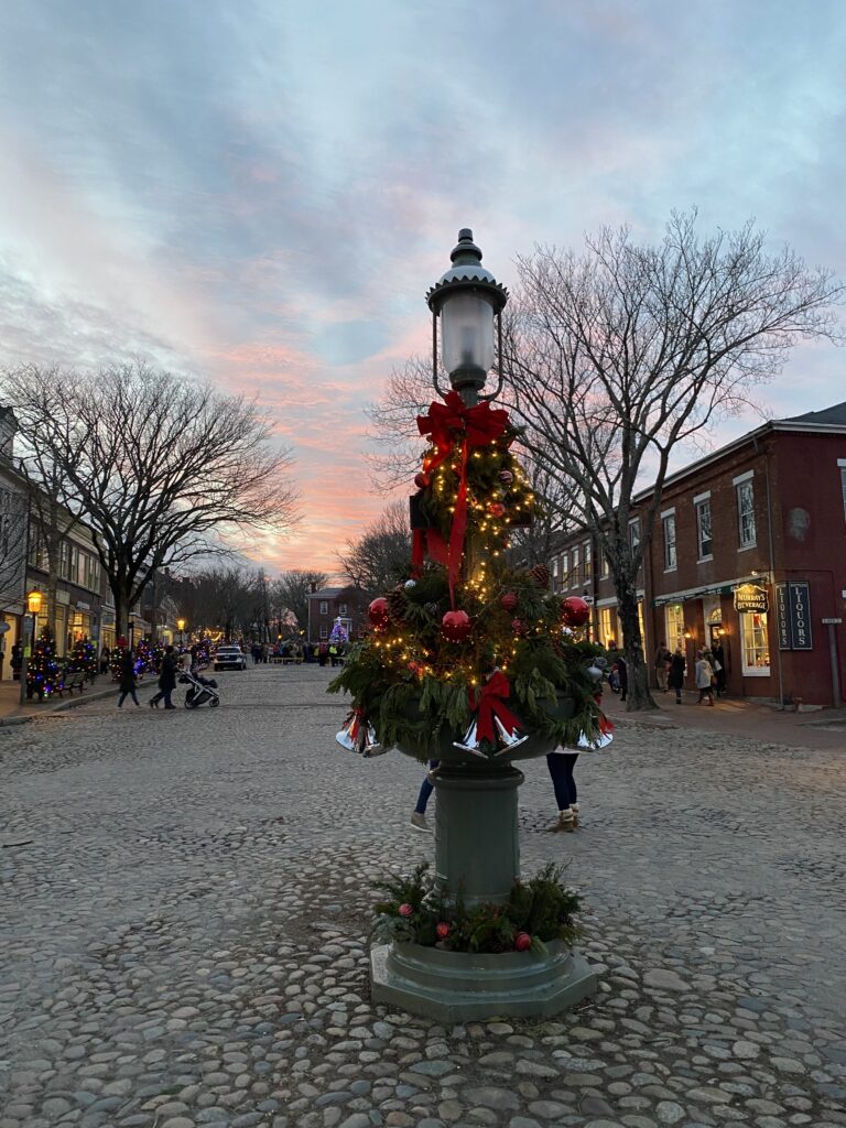 A decorated lamppost with greenery and lights stands on a cobblestone street at sunset, with people walking nearby.