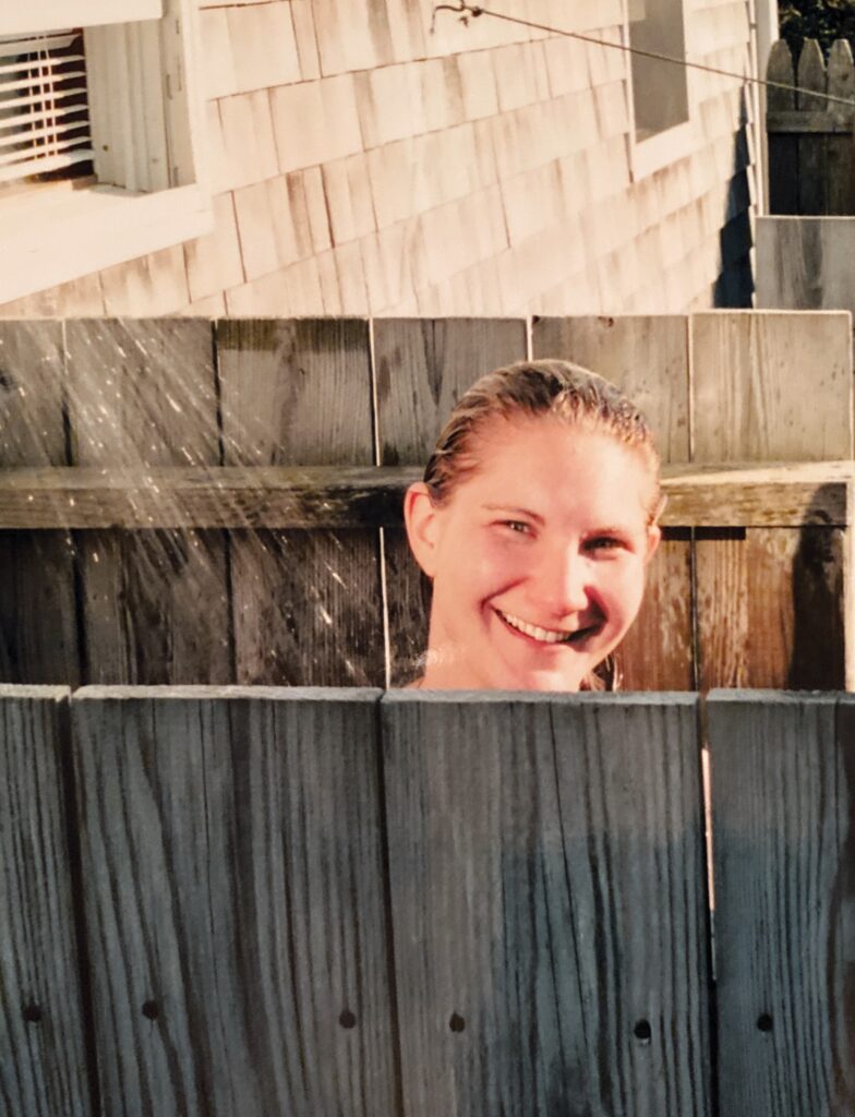 A smiling person with wet hair peeks over a wooden fence while water sprays around them.