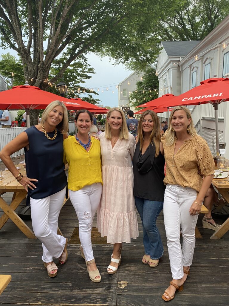 Five women pose together outdoors, smiling, wearing colorful outfits, with red umbrellas and trees in the background.