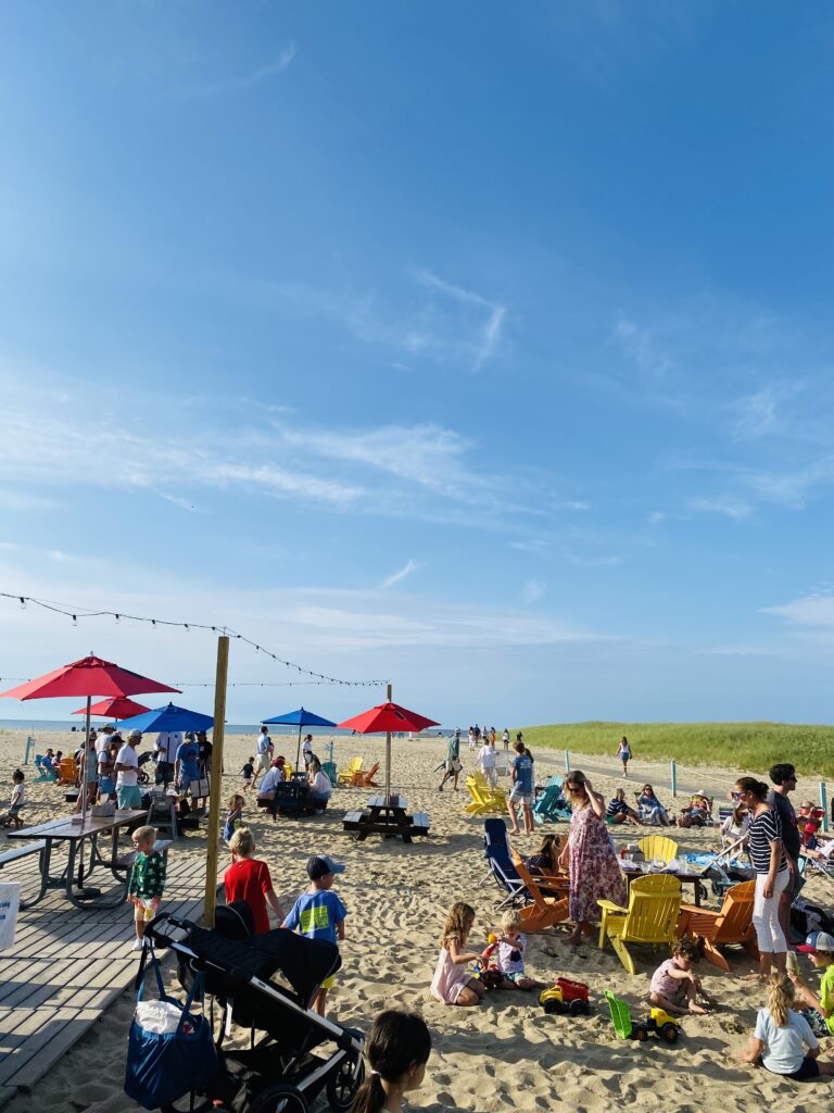 Crowd of people enjoying a sunny beach day with colorful chairs, children playing, and umbrellas in the background.