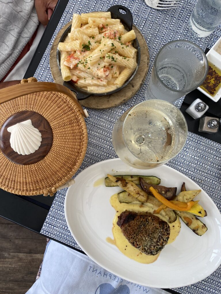 A plate with rigatoni pasta, a steak with sauce, and grilled vegetables, alongside drinks and a woven basket.