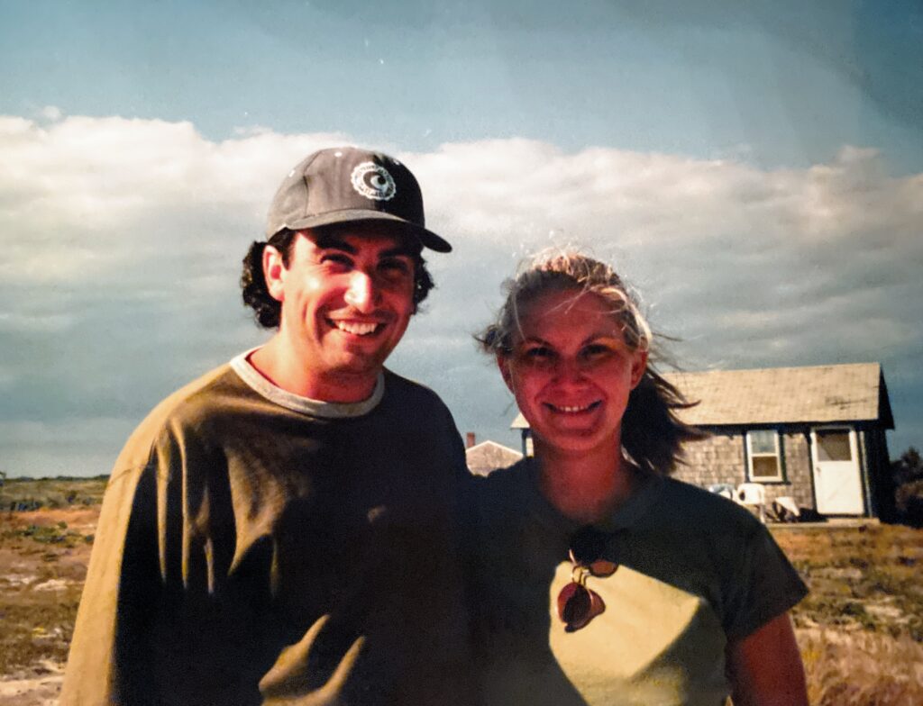 Two smiling individuals stand outdoors, with a small house in the background and a cloudy sky above.