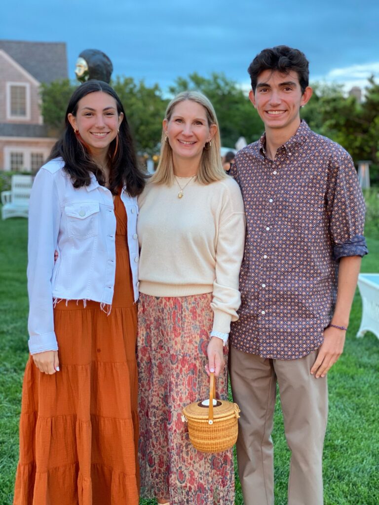 Three people pose together outdoors, smiling. One holds a small woven basket. Green grass and trees in the background.