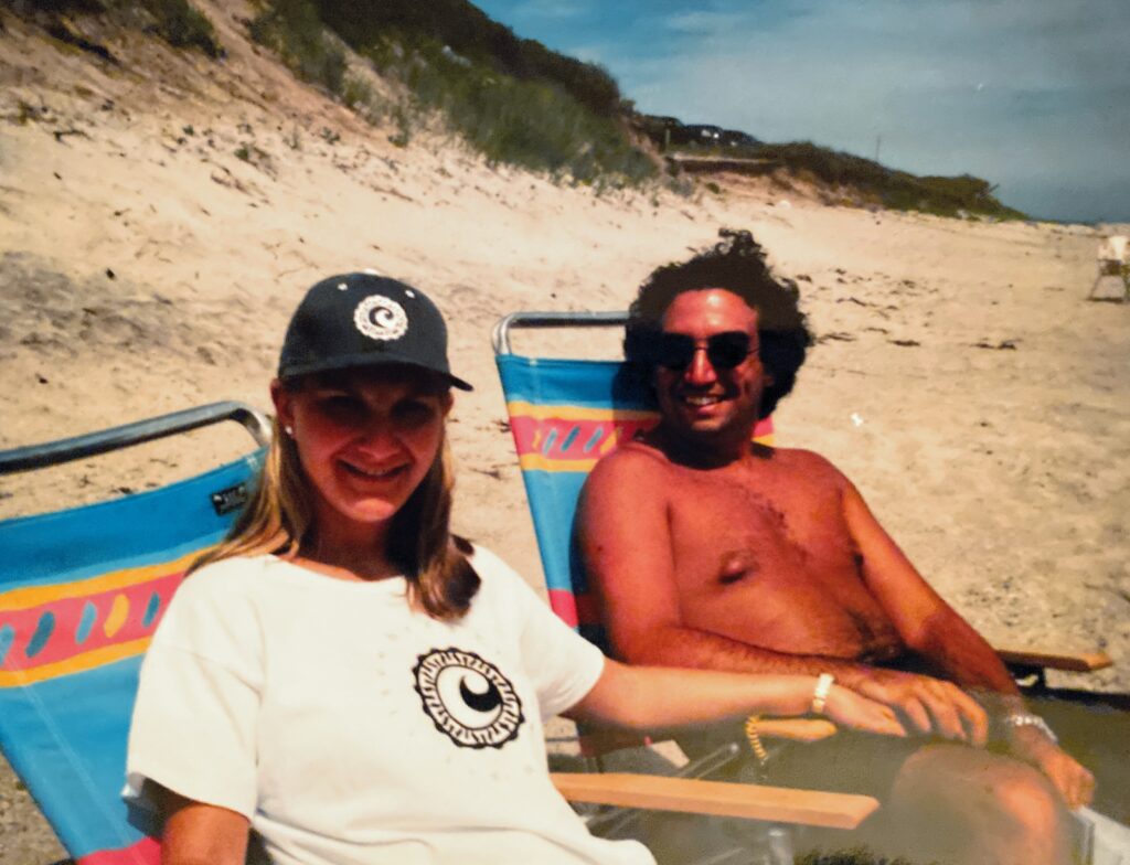 Two people relax on beach chairs, smiling at the camera, with sand and greenery in the background.