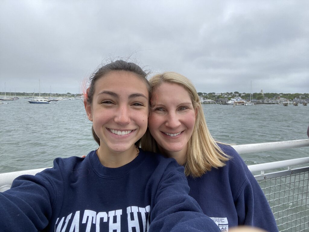 Two women smile for a selfie on a dock, with boats and a cloudy sky in the background.