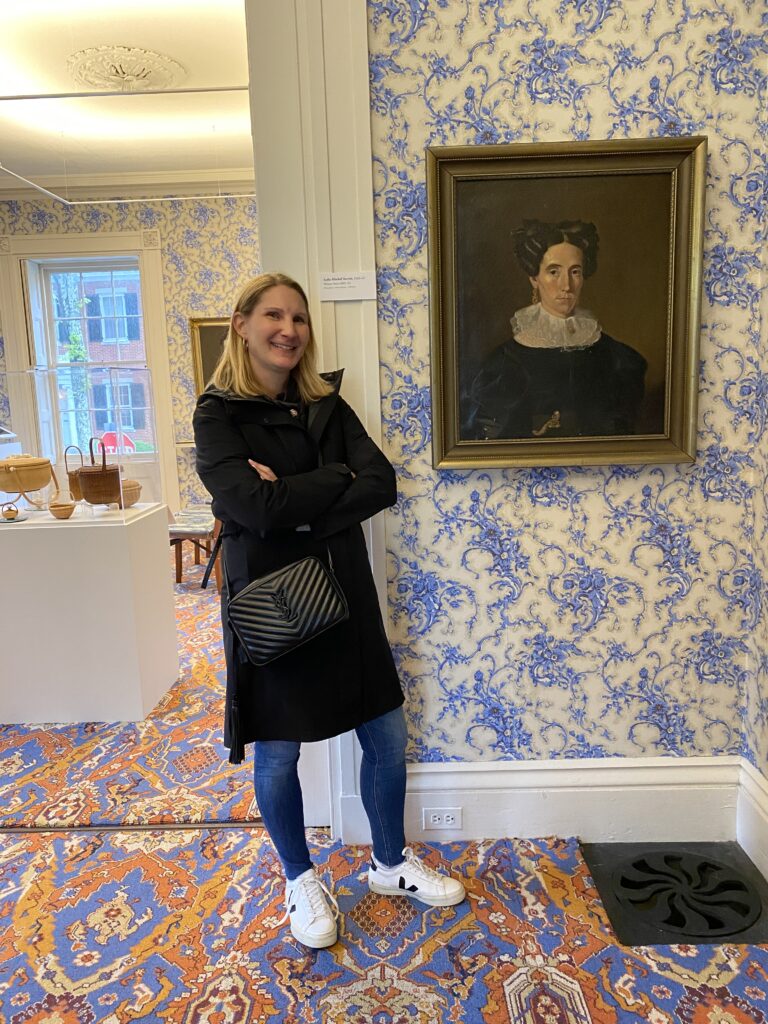 A woman stands beside a portrait in a patterned room, smiling with arms crossed.