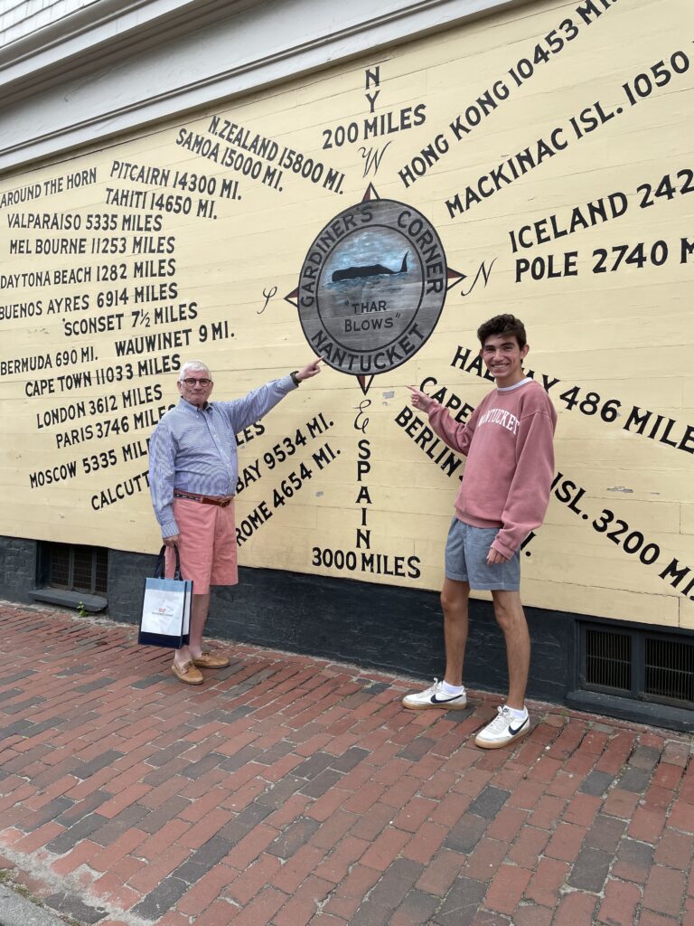 Two men stand in front of a wall mural displaying distances to various global locations.