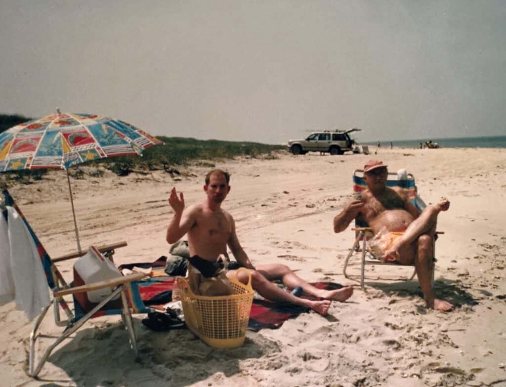 Two men relax on a beach, one waves while the other holds food. An umbrella and vehicle are in the background.