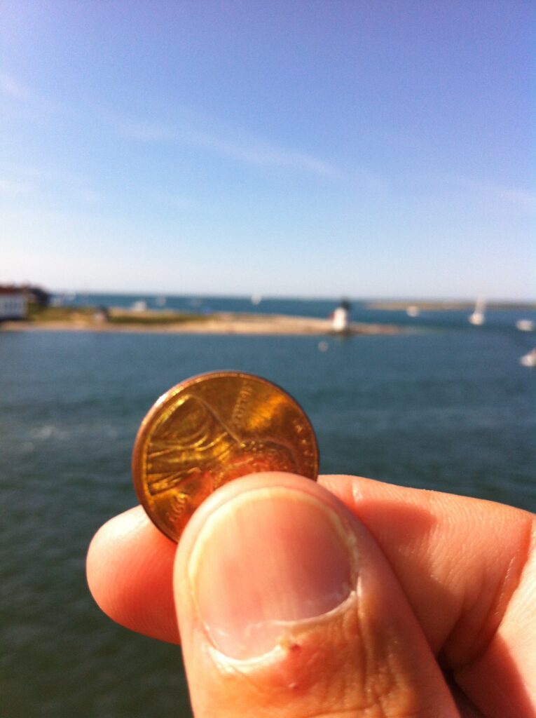 A person holds a penny in front of a coastal scene with a lighthouse and boats in the background.