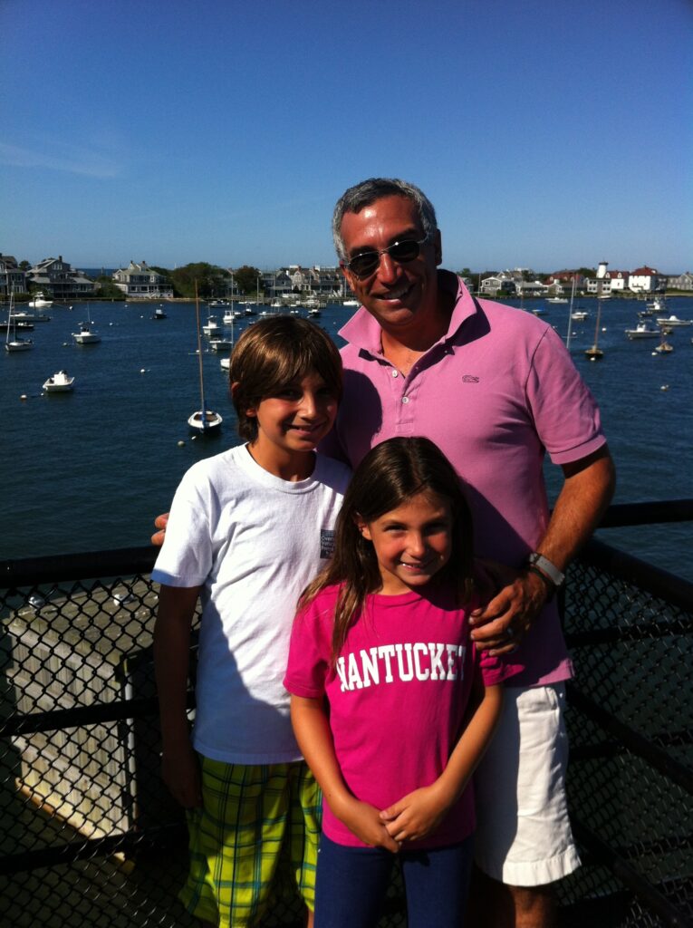 Three people pose together by a waterfront, smiling. A man stands behind two children, with boats in the background.