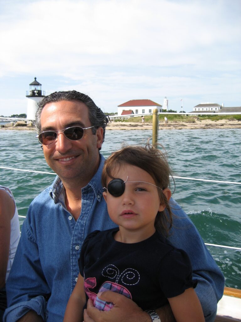 A man smiles while holding a girl wearing an eye patch, with a lighthouse and buildings in the background.