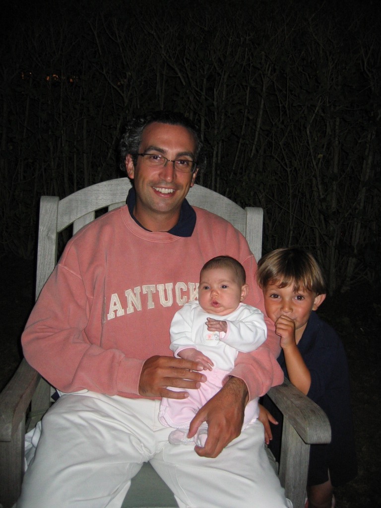 A man sits on a chair holding a baby, while a young boy stands beside them, smiling.
