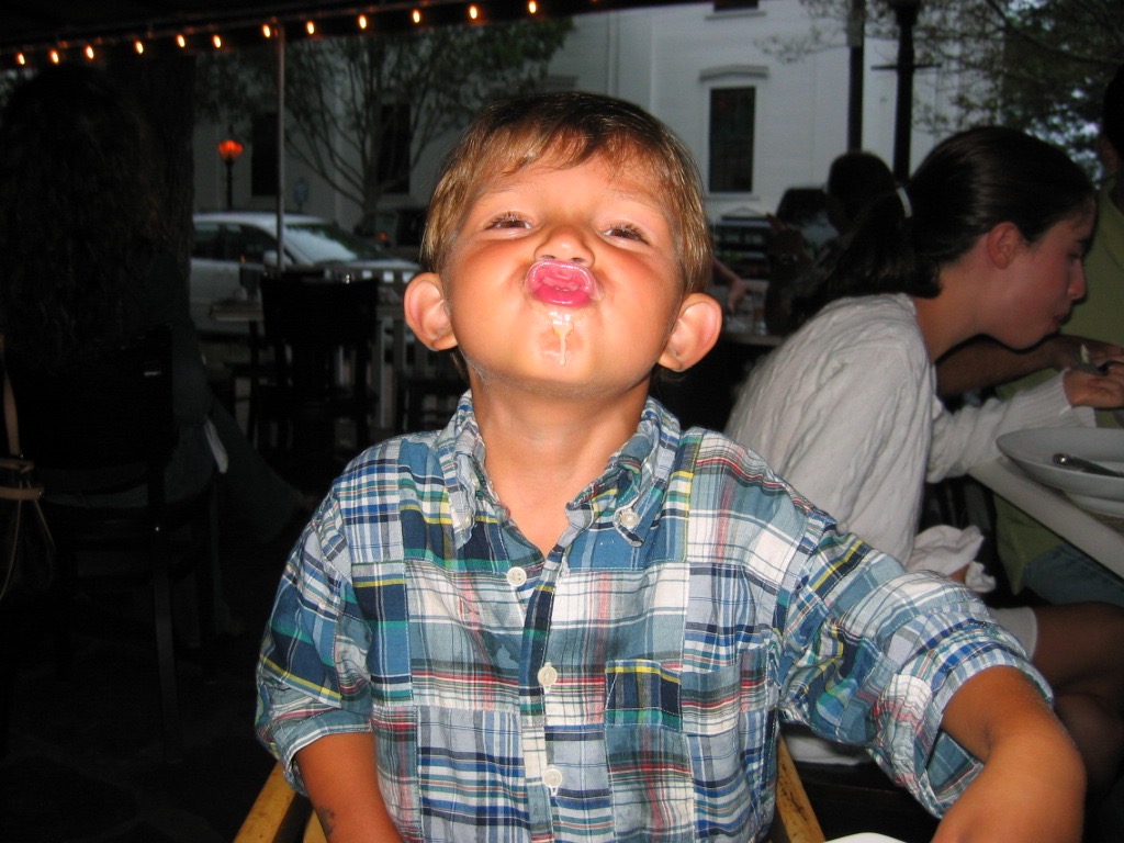 A young boy in a plaid shirt makes a funny face, with food on his lips, sitting at a restaurant table.