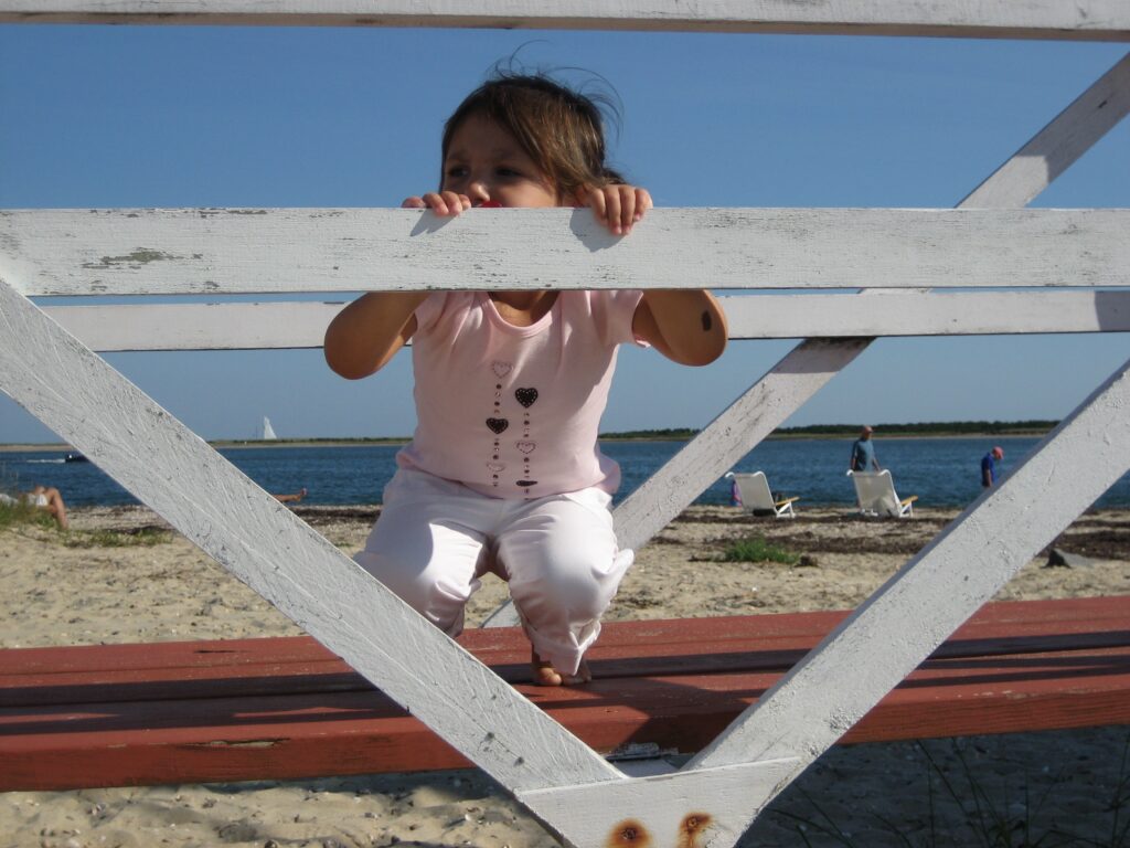 A child in a pink shirt climbs on a wooden structure near the beach, with water and people in the background.