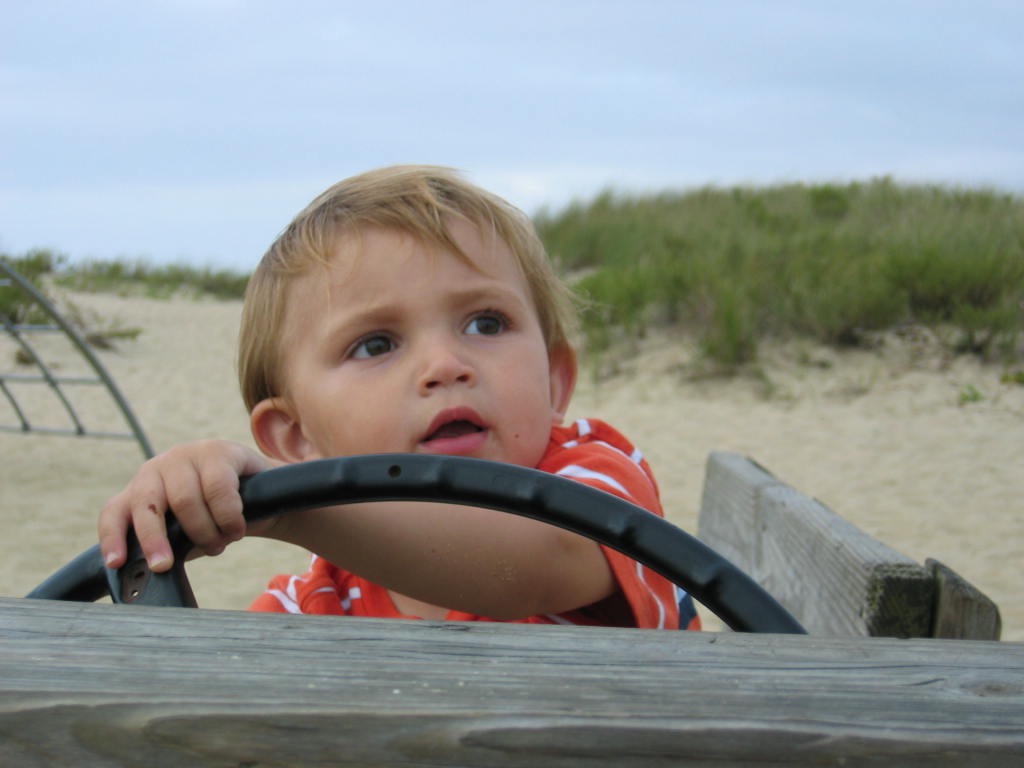 A young child with short hair grips a steering wheel, looking thoughtfully into the distance at a sandy playground.