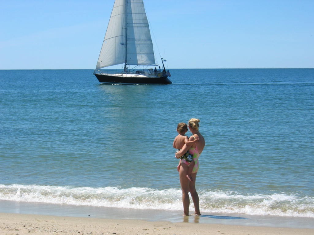 A woman holds a child on the beach, with a sailboat in the background and gentle waves lapping at the shore.
