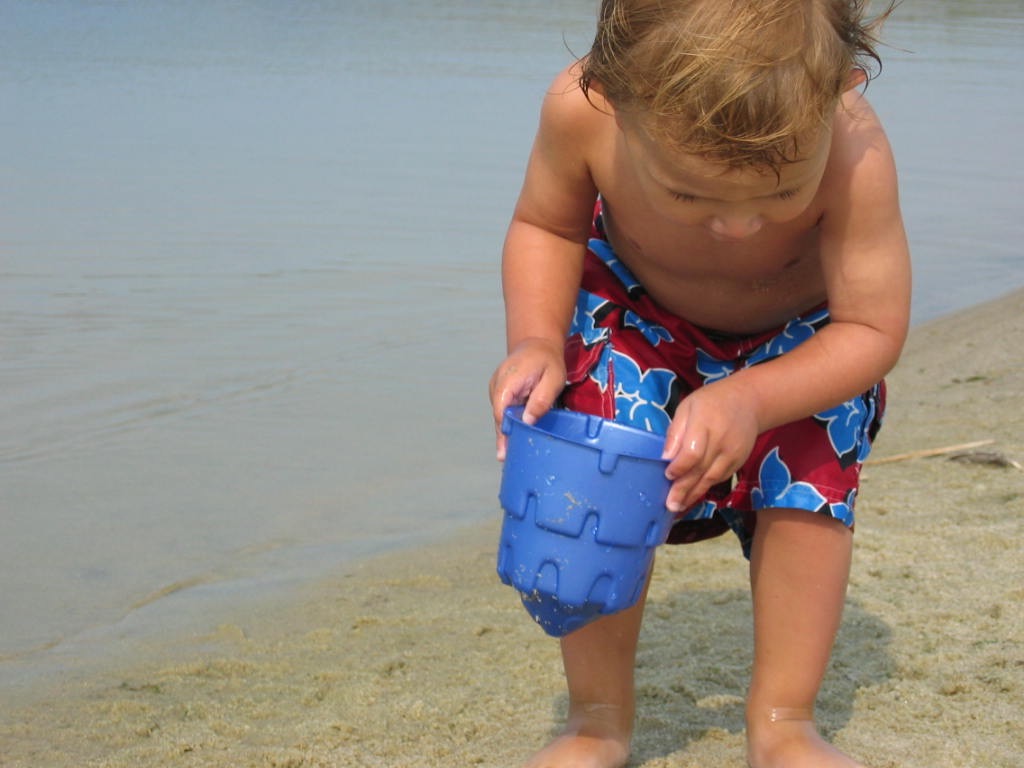 A child in swim trunks scoops sand with a blue bucket at the water's edge.