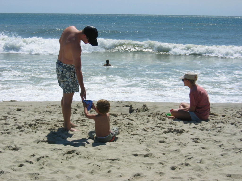 A man helps a child with a sand toy on the beach while a woman sits nearby, watching the ocean.