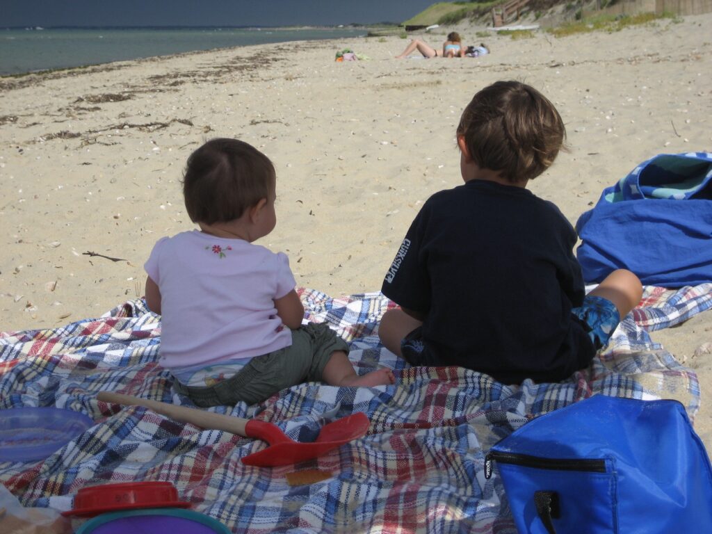 Two children sit on a blanket at the beach, facing the ocean, with toys scattered around them.