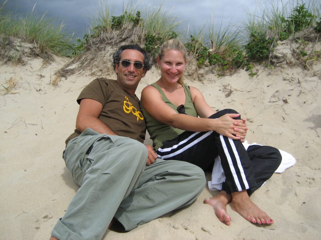 Two people sit on a sandy beach, smiling and relaxed, with grass-covered dunes in the background.
