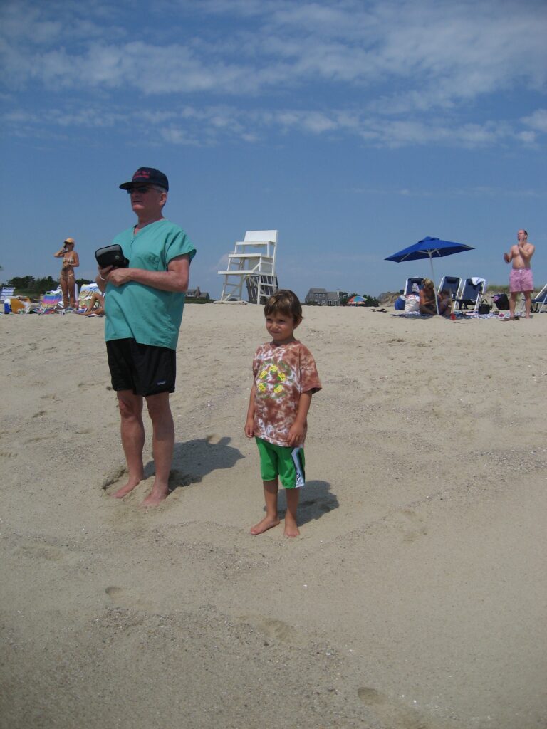 An adult man and a child stand on the beach, with beachgoers and a lifeguard chair in the background.