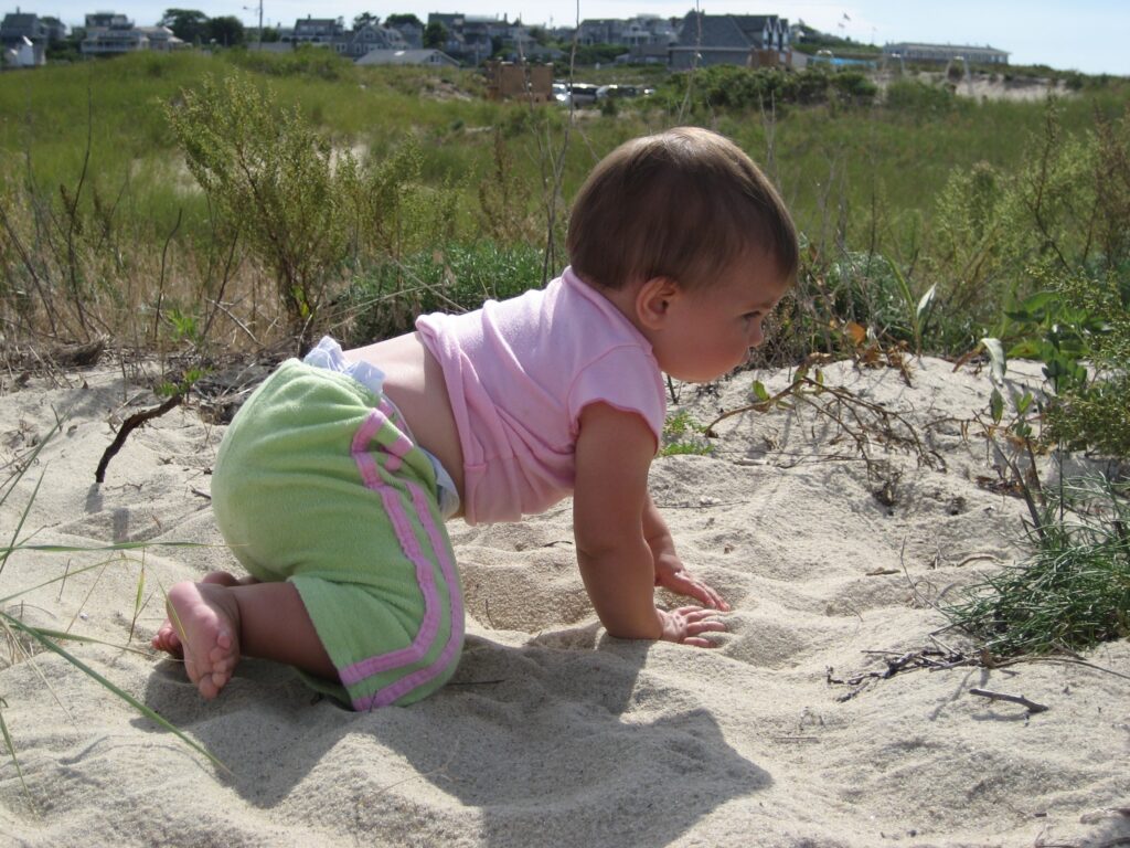 A baby crawls on sandy ground, focused on the surroundings, with grass and houses in the background.
