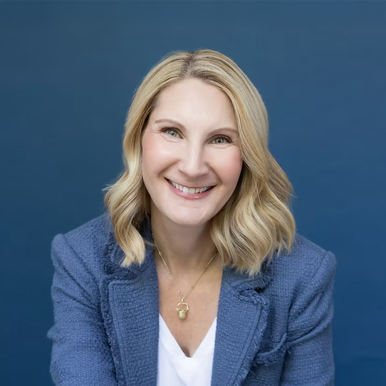 A woman with wavy blonde hair smiles, wearing a blue blazer against a blue background.